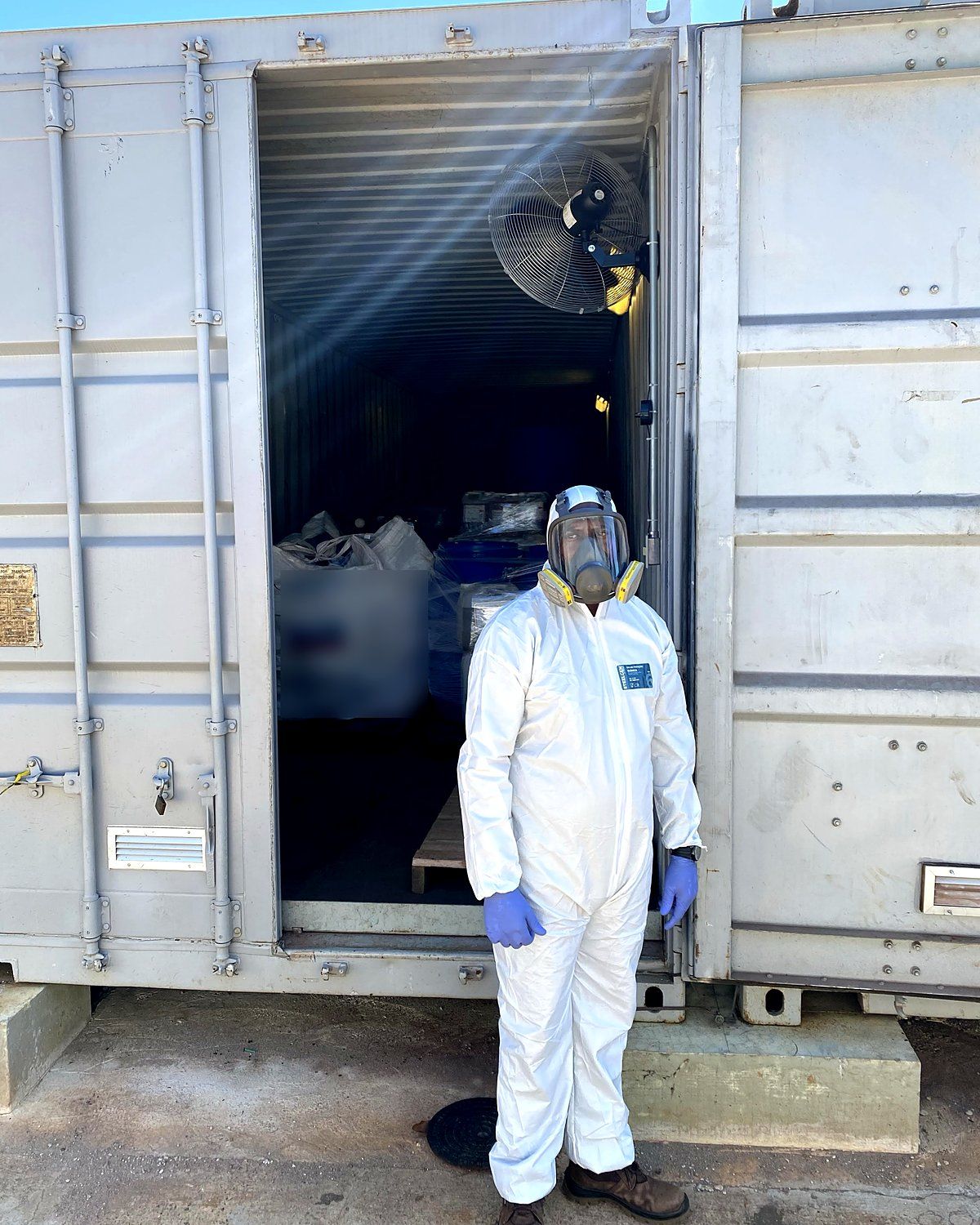 Operador PGRS com macacão de proteção química, respirador facial e luvas em frente a contêiner de armazenamento